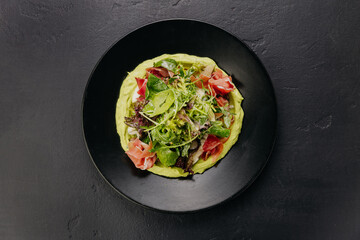 Fresh salad with avocado cream and prosciutto, served on a black plate, perfect for gourmet dining