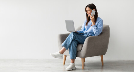 Full length of Caucasian lady using laptop and speaking on smartphone while sitting in armchair...