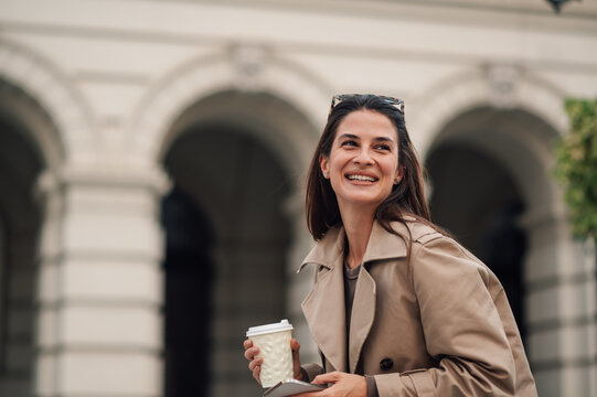 Smiling businesswoman holding coffee and smartphone in urban city center - Powered by Adobe