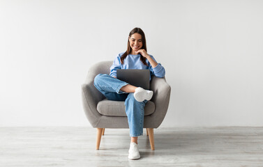 Millennial Caucasian lady in casual wear using laptop, sitting in armchair, having online...