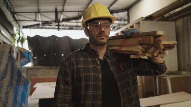 A handsome professional Caucasian male carpenter worker wearing a yellow helmet and safety goggles is working and walking carrying timber on his shoulder at his workplace in the furniture factory. 