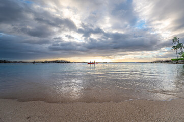 Obraz premium As the sun sets, two individuals paddle in a kayak on a peaceful beach while dark clouds loom overhead, reflecting the day's last light on the water's surface.