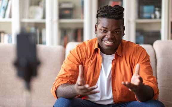Blogging concept. Young African American chuby guy blogger in casual clothes streaming video online or recording tutorial for social media network, looking at the mobile phone on a tripod and smiling