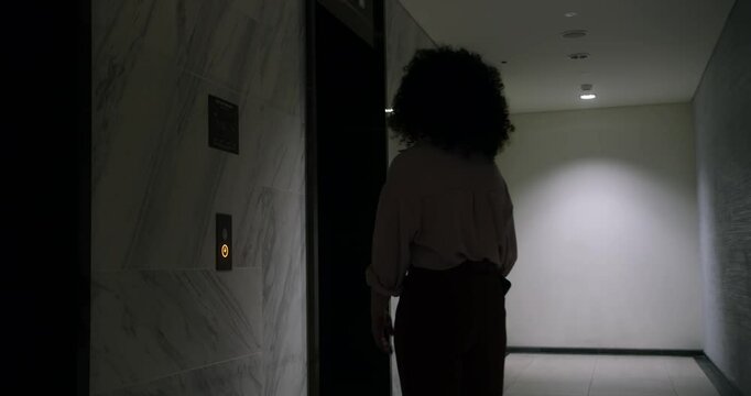 A businesswoman presses the elevator call button and waits for the elevator. A woman with curly hair wearing glasses with tablet enters an elevator.