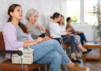 Young girl in casual clothes sitting in lobby of medical clinic, waiting for appointment with...