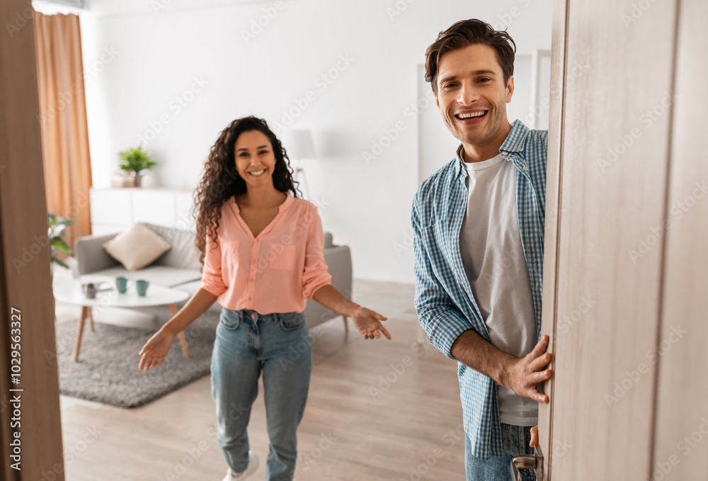 Wall mural Portrait of cheerful couple welcoming inviting guests to enter home, happy young guy and lady standing in doorway of modern flat, looking out together, waiting for visitor friends to come in