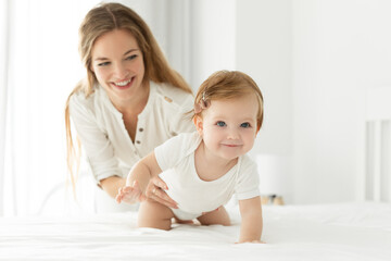 Sweet baby girl in white bodysuit crawling on bed while mother looking at daughter on background. Bright home setting showing family bonding moments