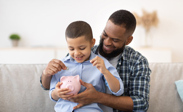 Financial Literacy. Cheerful African Father And Son Putting Money In Piggybank Sitting On Sofa At Home. Daddy Teaching His Child Budget Planning, Keeping Personal Savings Safety