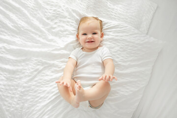 Pretty smiling baby girl playing with her legs lying on her back on white cotton bed in the bedroom and looking at camera, top view. Care for child concept