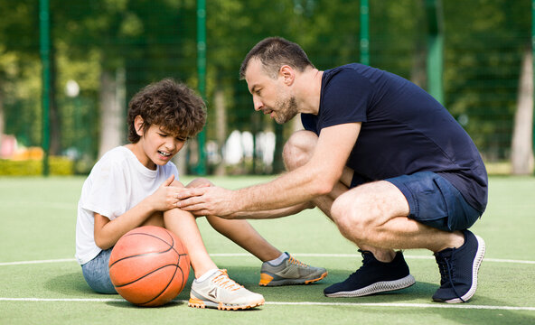 Sports Injury. Handsome PE teacher helping boy with knee trauma after playing basketball