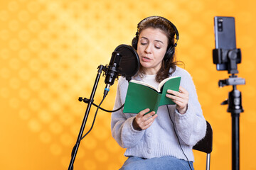 Influencer filming herself reading book, creating audiobook, isolated over studio background. Woman...