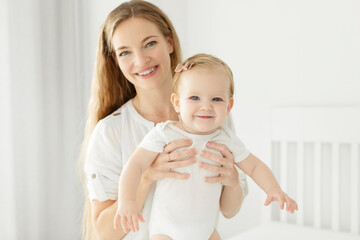 Maternal love and care concept. Beautiful European mother with baby girl having fun on the bed in bright bedroom, happy motherhood with maternity leave