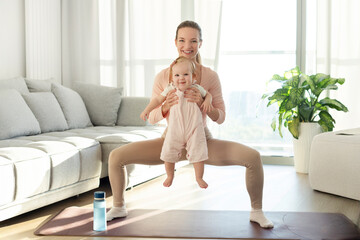 Obraz premium Young woman fit mom with baby girl in her arms doing fitness, squat exercise on mat at home, training with child
