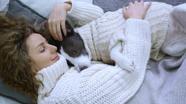 Woman in sweater hugs dog on soft bed. Gently scratching pet, woman radiates warmth and love. Dog in sweater looks happy. Woman and four-legged friend create atmosphere comfort and coziness in bedroom