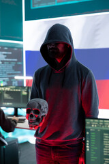 Digital outlaw hacker in a dark hood issuing cyber threats with a human skull to make death...