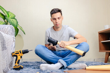 A man is tapping a wooden leg with a rubber mallet to assemble a small table. In the background, there is a scene of a small living room. DIY furniture assembly