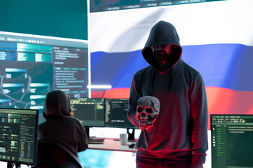 Conceptual image of a hooded russian hacker creating hazardous alerts, holds a skull and warning of death threats. Russian federation works on cybercrime and violence, panic and torment.