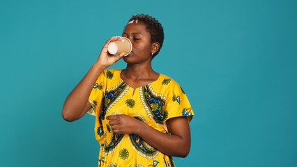 Young smiling person enjoying freshness of coffee cup in studio, drinking caffeine refreshment and relaxing against blue background. Happy woman serving a fresh beverage. Camera A.