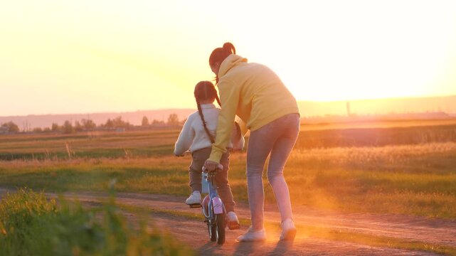 Careful mother holds little girl to learn bicycling in sunset park. Little girl with mother learns to ride bicycle in evening rural park. Mother and girl play together with bicycle in spring meadow