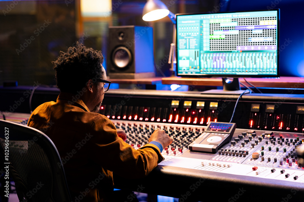 Poster African american sound designer uses mixing console with knobs and faders in control room, editing old recordings and adding effects. Skilled audio expert producing music at mixer board.
