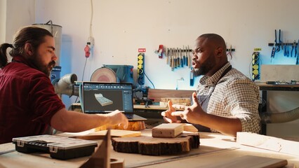Carpenter looking over blueprints schematics on laptop in CAD software with apprentice. Woodworkers...