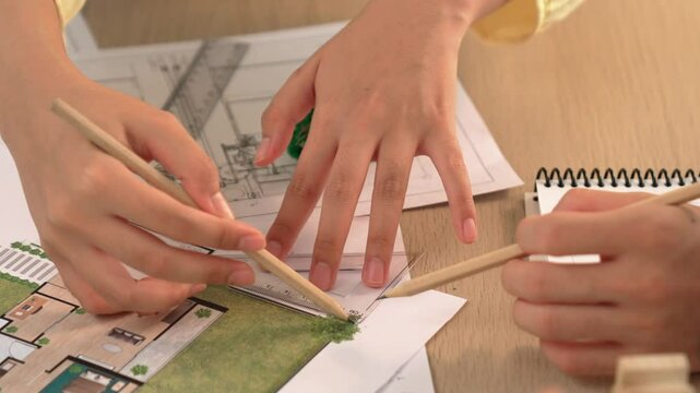 Architects using measurement tool and pencil tracing in building blueprint by coworker support on table with architectural document and wooden block scatter around at office. Closeup. Delineation.