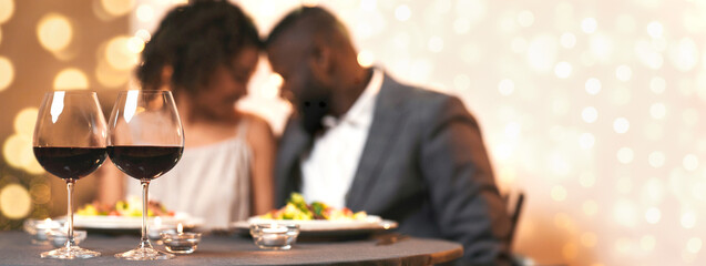 Romantic dinner concept. Two glasses of red wine on festive dinner table over cuddling afro couple