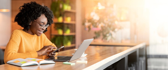 Happy black girl using mobile phone, working with laptop in cafe, side view, copy space