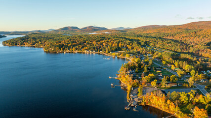 A stunning sunrise over Schroon Lake, NY, with warm light casting a soft glow on the calm water and surrounding autumn-colored forests and mountains.