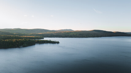 A stunning sunrise over Schroon Lake, NY, with warm light casting a soft glow on the calm water and surrounding autumn-colored forests and mountains.
