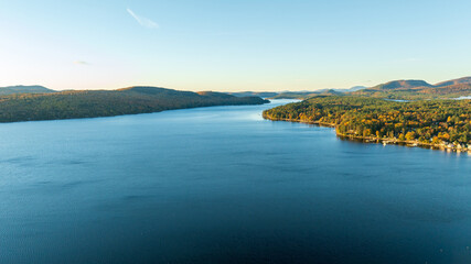 A stunning sunrise over Schroon Lake, NY, with warm light casting a soft glow on the calm water and surrounding autumn-colored forests and mountains.