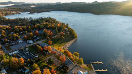 A stunning sunrise over Schroon Lake, NY, with warm light casting a soft glow on the calm water and surrounding autumn-colored forests and mountains.