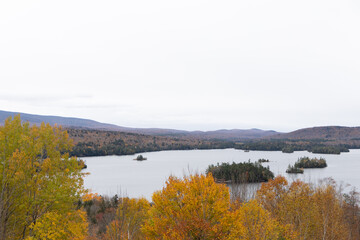 Obraz premium Scenic view of Blue Mountain Lake, NY, with autumn foliage surrounding the lake's islands and rolling hills in the background, showcasing the vibrant beauty of fall in the Adirondacks.
