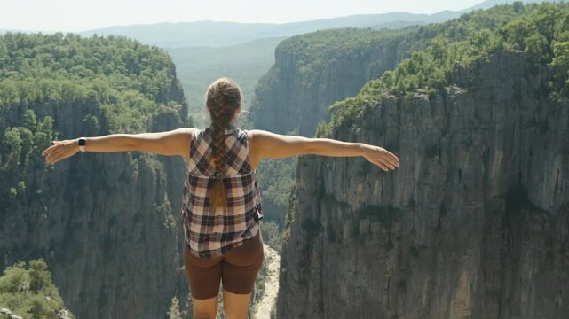 A close-up view of a Woman standing on a cliff with her arms stretched out, facing the vast Canyon. The green forest and mountainous landscape stretch far into the horizon.