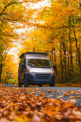 Low Angle Camper Van Drives Remote Forest Road Off the Grid Leaves on Ground in Autumn Season Fall Foliage. Headlights On RV on Street USA Scenic