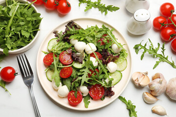 Tasty salad with arugula, lettuce, mozzarella cheese, vegetables and fork on white table, flat lay