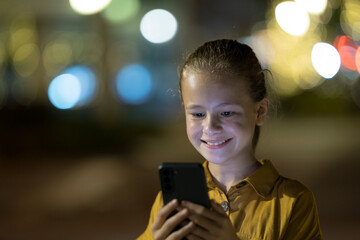 Positive child girl messaging on her mobile phone with a friend outdoors at night