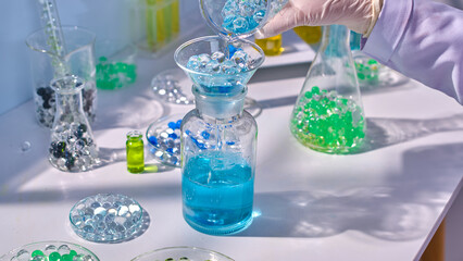 Blue essence in glass beaker poured into glass bottle on table through a funnel, some lab glassware contain colorful gel balls placed around. High angle view, empty space for adding elements of design