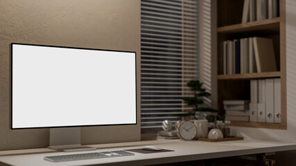 A computer with a white screen mockup on a table against the wall in a minimalist office at night.