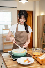 A happy Asian woman is cooking in the kitchen, making scrambled egg in a pan for her breakfast.