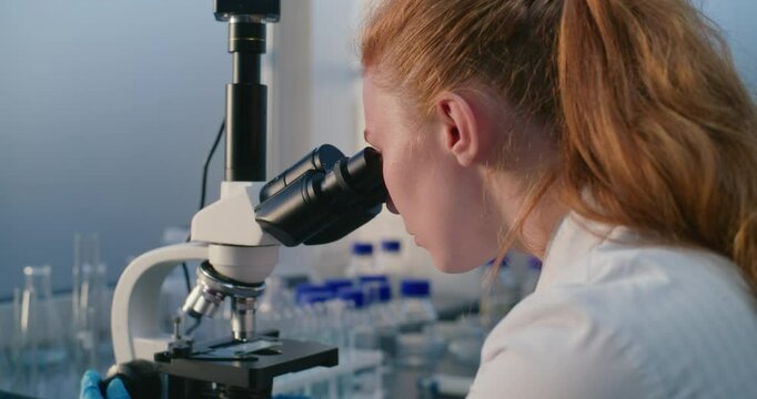 Female scientist, chemist sits and works with microscope in bright medical science laboratory. Biochemistry does analysis of new drug sample. Development of pharmacology. Pharmaceuticals and medicine.