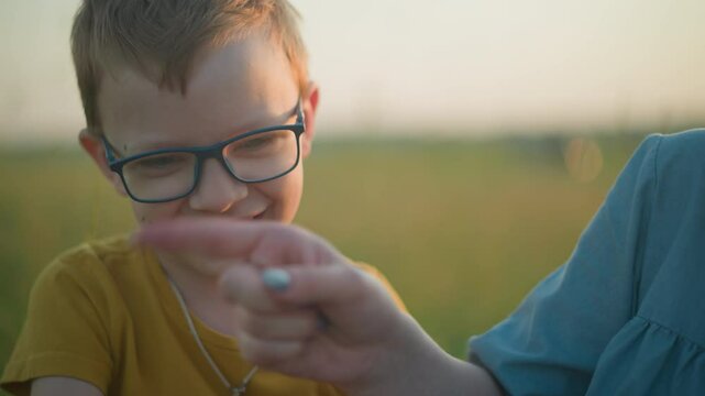 A young boy dressed in yellow playfully distracts his partially visible mother, who is sitting beside him in a grassy field. The boy smiles mischievously, enjoying the interaction with his mom
