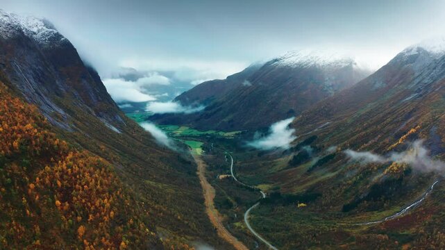 A scenic aerial view captures a fog-filled mountain valley, with vibrant fall foliage, a river, and a road below. A glacier lies at the end of the valley.