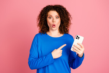 Young woman in blue sweatshirt expressing amazement while holding smartphone against pink background