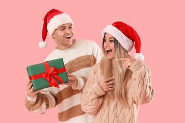 Happy young couple in Santa hats with Christmas gift on pink background