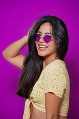 Woman with purple sunglasses and long hair, wearing a yellow floral top, smiling confidently in a trendy, playful pose against a vibrant backdrop. 