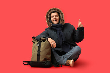 Young man in stylish puffer jacket and scarf with bag pointing at something on red background