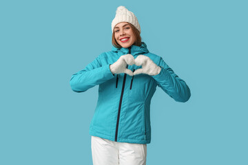 Beautiful young happy woman in warm winter clothes showing heart gesture on blue background