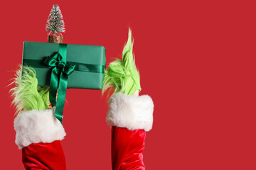Green hairy hands of creature in Santa costume with decorative fir tree and Christmas gift box pointing at something on red background