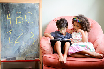Two children sitting together on a pink chair, using a tablet, w